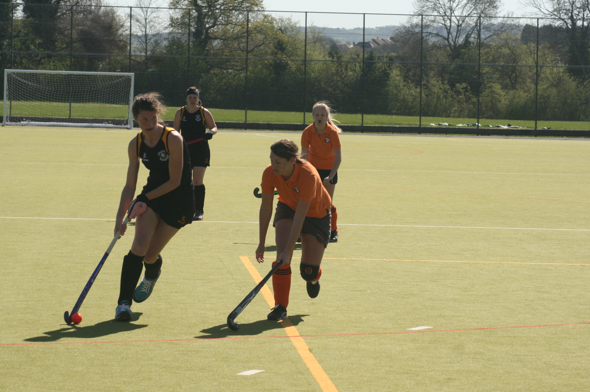 A field hockey player in a black kit dribbling the ball while an opponent in an orange kit challenges for it